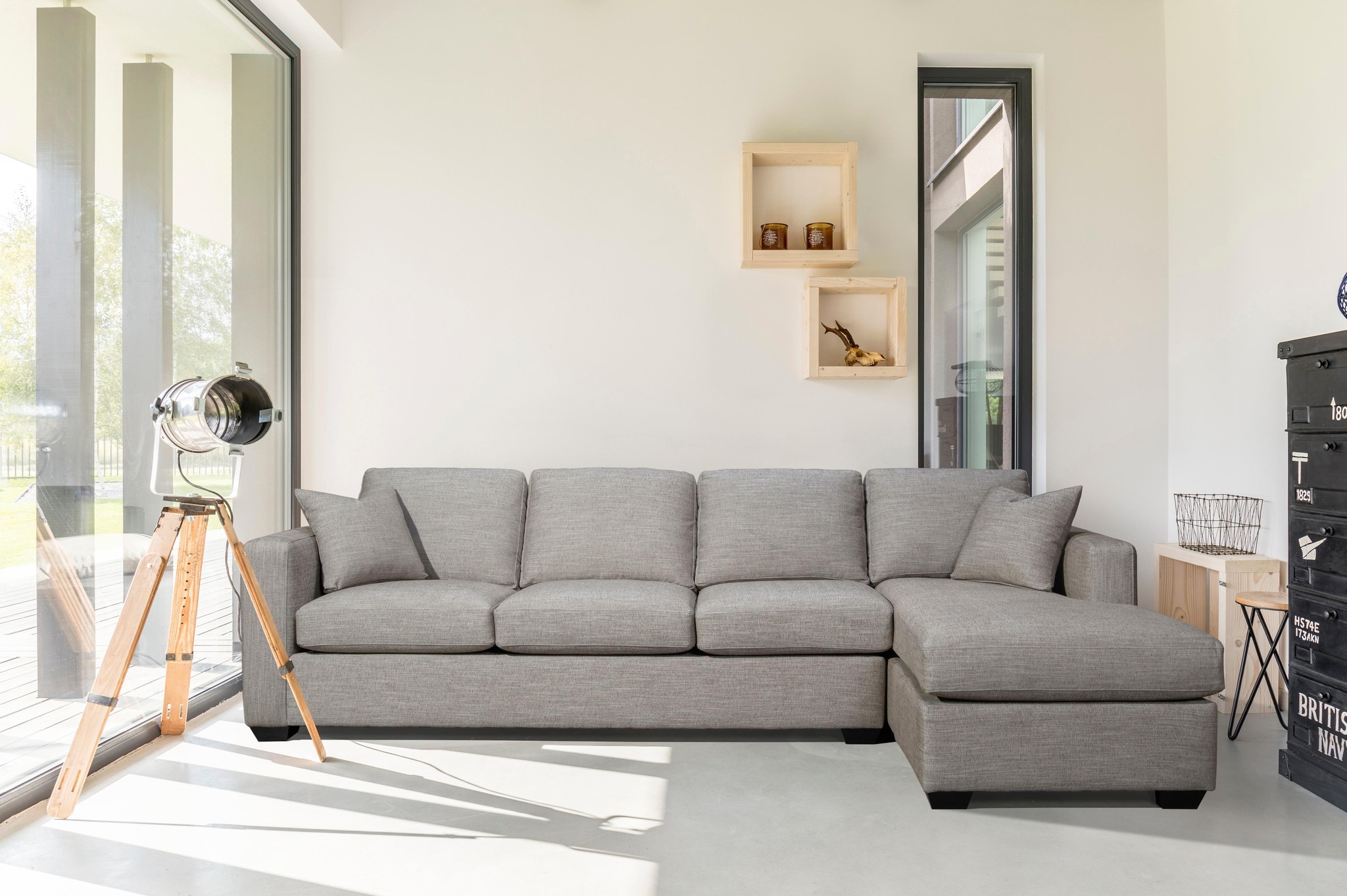 Salon contemporain avec un grand canapé d’angle gris, une lampe sur trépied en bois, des étagères murales minimalistes et une décoration industrielle, baigné de lumière naturelle de chez  ​Ameublement BrandSource S.J. Dion