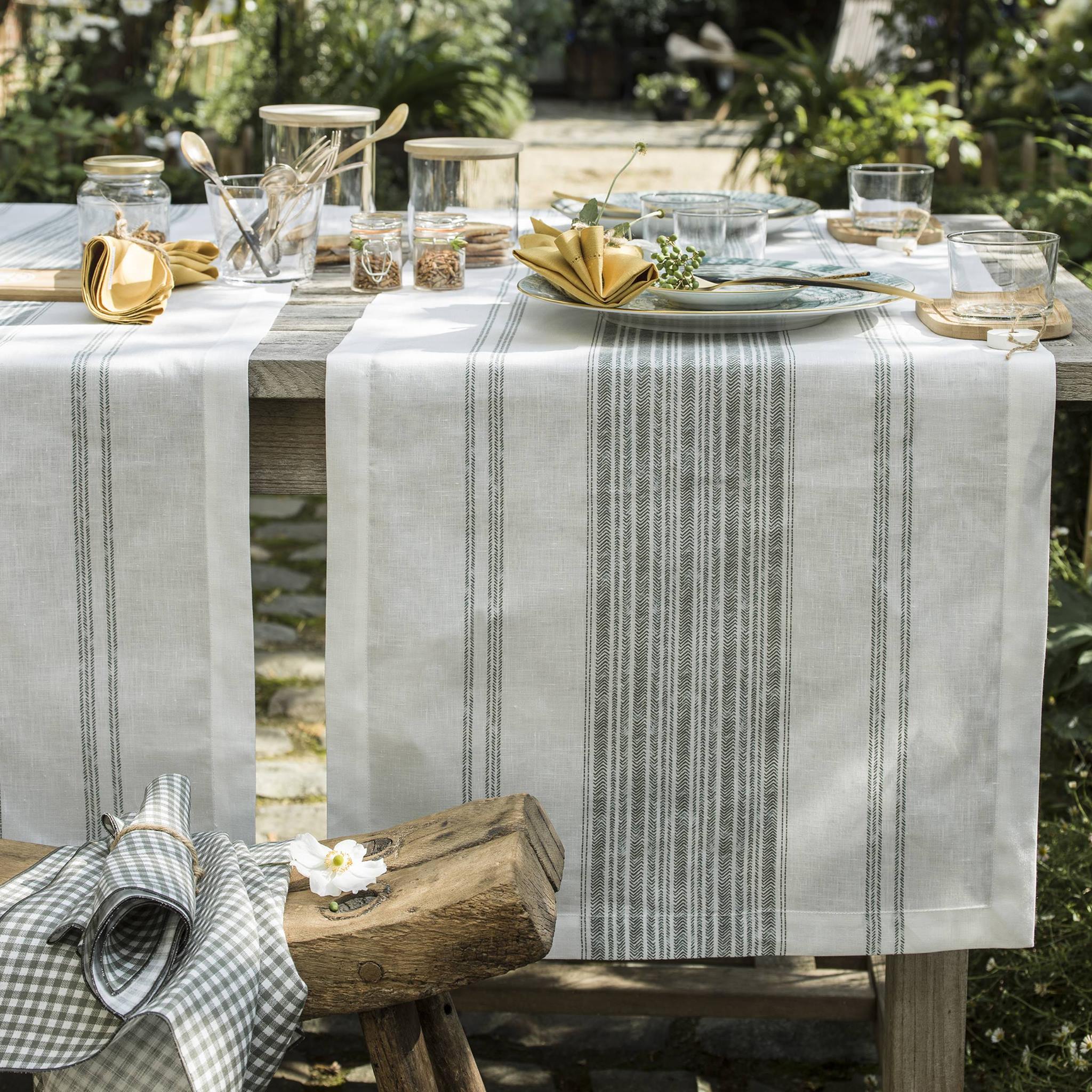 Table en bois dressée en plein air avec chemins de table en lin à motifs rayés, vaisselle élégante et serviettes jaunes pliées, entourée de verdure de chez AU LOFT.