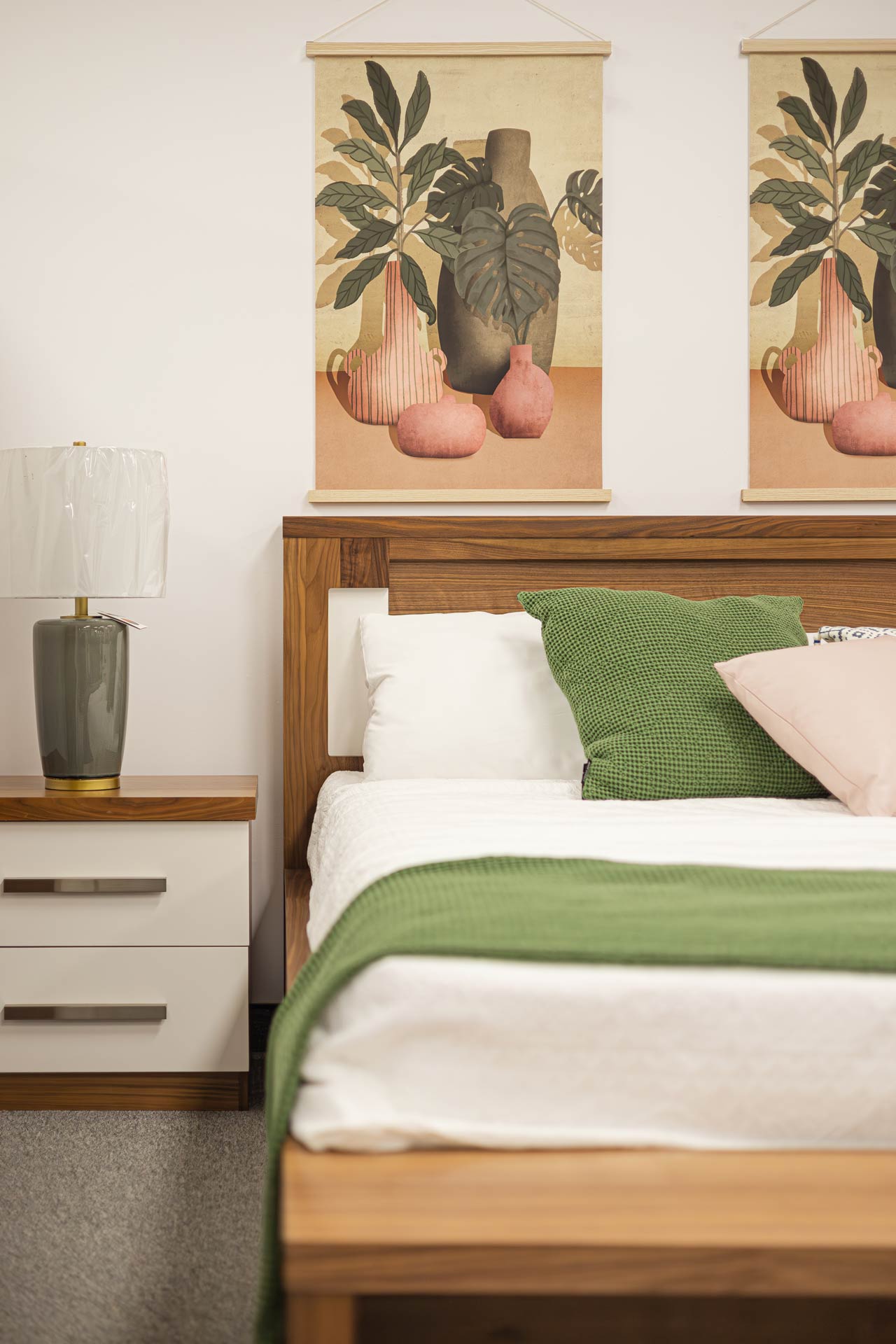 Chambre à coucher moderne avec un lit en bois, des coussins verts et roses, une table de chevet blanche et une lampe élégante, accompagnée de décorations murales artistiques aux tons naturels de chez magasin Avivia
