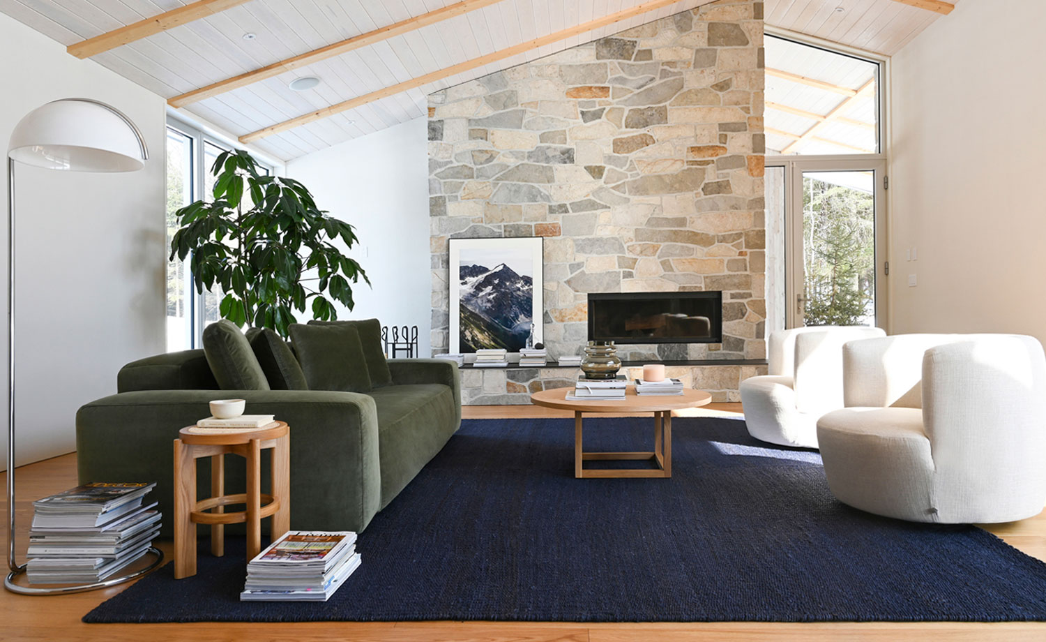 Salon lumineux au design moderne avec un canapé vert en velours, fauteuils arrondis blancs, tapis bleu profond et une cheminée en pierre naturelle, créant une ambiance chaleureuse et élégante de chez Élément de Base