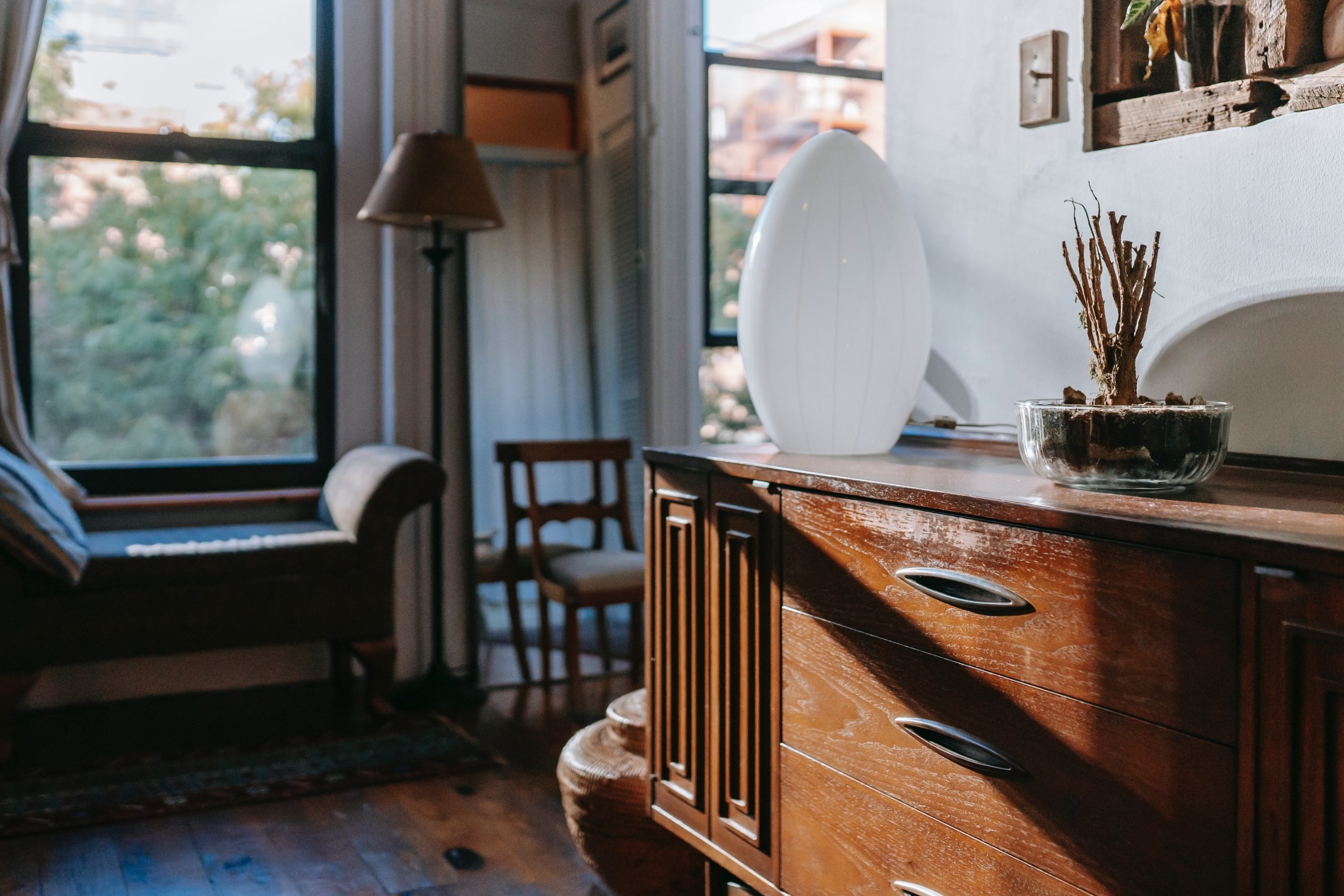 Buffet en bois vintage avec une lampe décorative blanche et une plante, dans un salon lumineux au style rétro avec vue sur l'extérieur.