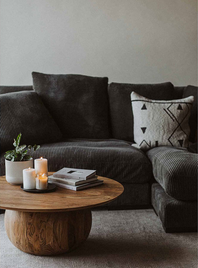Salon cosy avec un canapé en velours côtelé foncé, une table basse en bois massif décorée de bougies, de livres et d’une plante verte, créant une atmosphère apaisante et élégante de chez la Boutique michel Bourgeois Maison.
